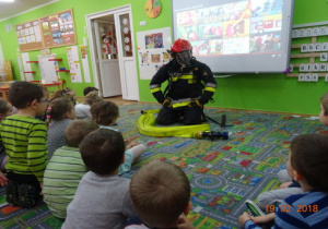 Strażak w umundurowaniu i kaskiem na głowie prezentuje dzieciom sprzet strazacki, w tle prezentacja multimedialna. Dzieci siedzą w półkolu na dywanie i uważnie słuchają. Przed strażakiem leży wąż strażacki.